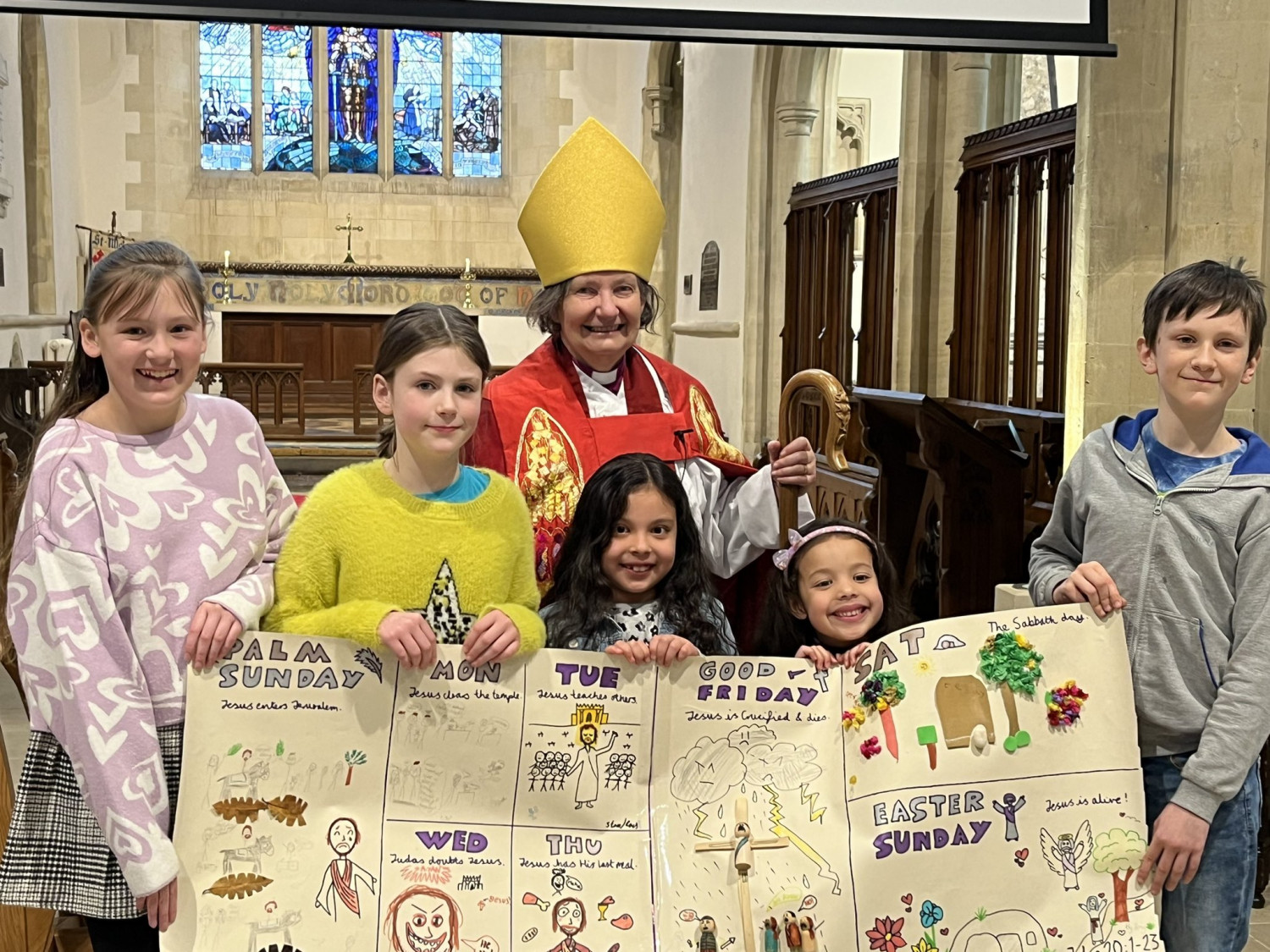 Photo of Bishop Viv standing behind 5 children of differing ages who are holding up a large poster with comic style art of the Easter calendar.